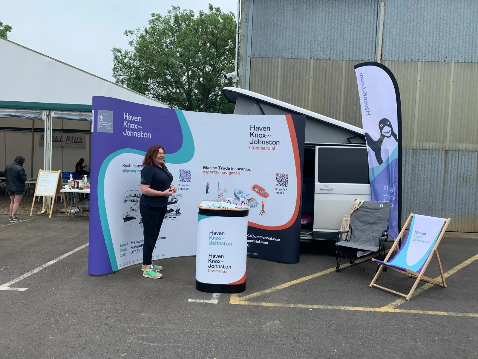 A woman stands at a Haven Knox-Johnston Commercial booth with banners, a table, and a van in the background, set up in an outdoor area.