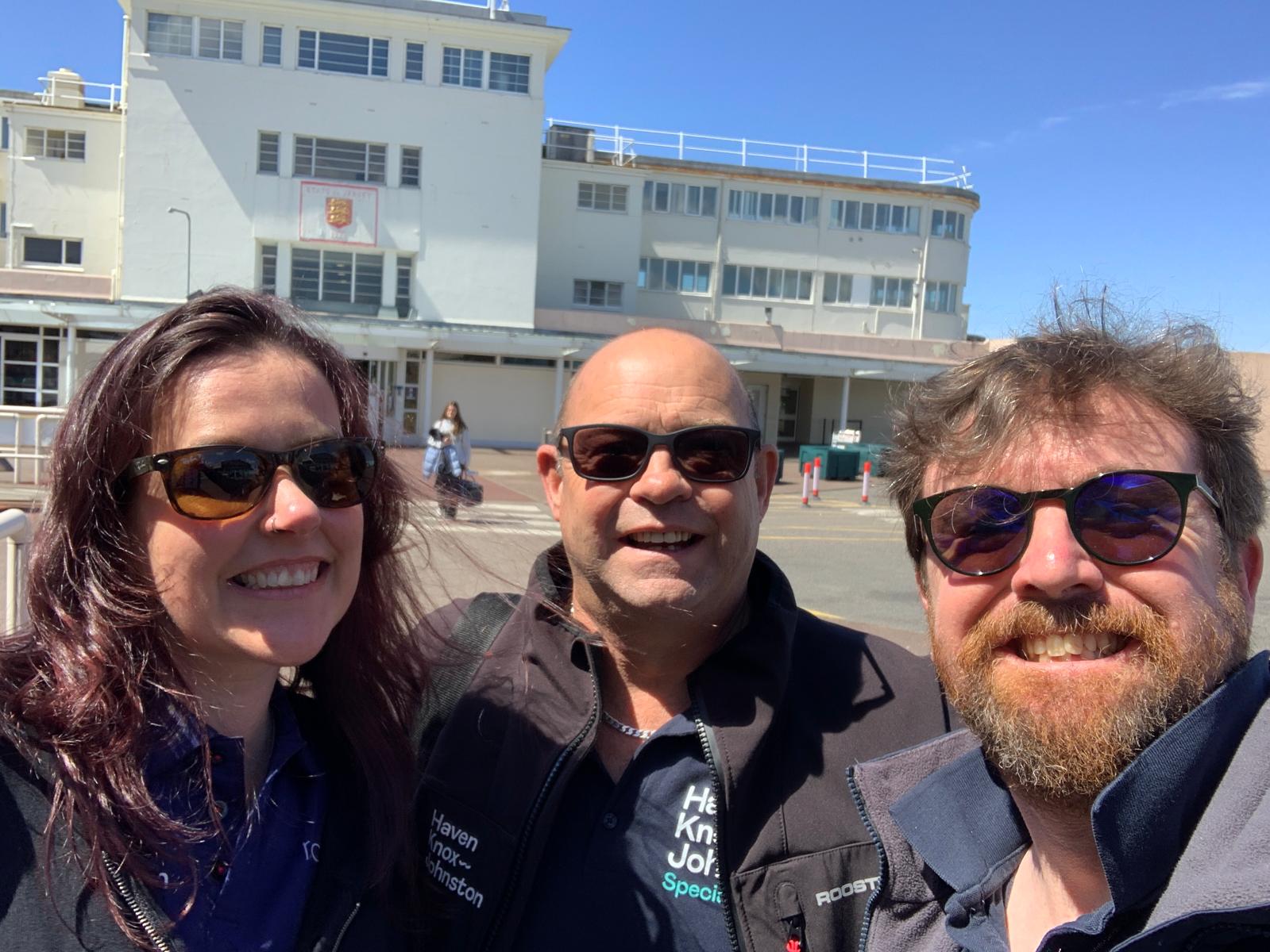 Three people wearing sunglasses smile for a photo outdoors, with a large building and clear sky in the background.