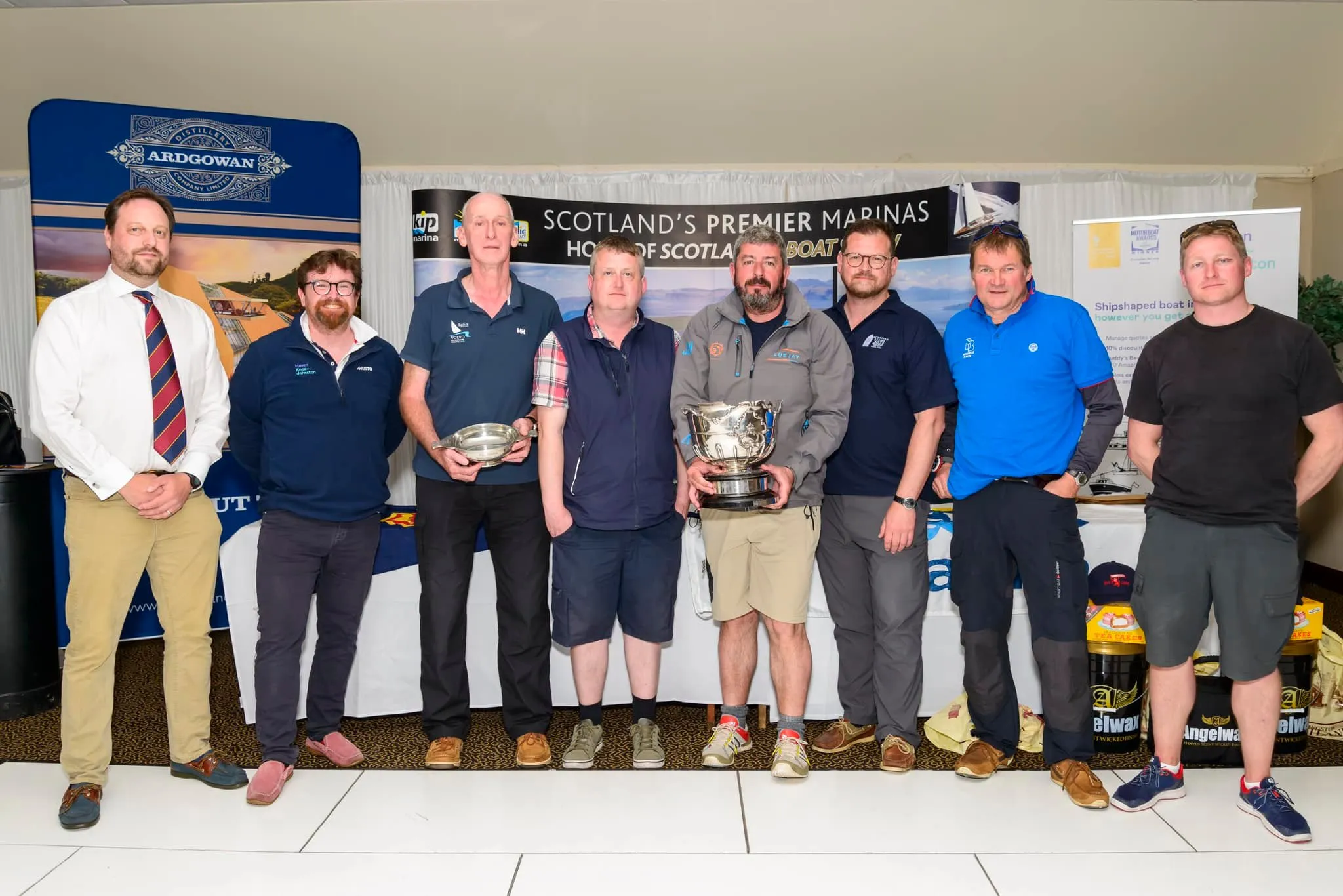 A group of eight men stand in a row indoors, several holding trophies, in front of a banner for Scotland's Premier Marinas.