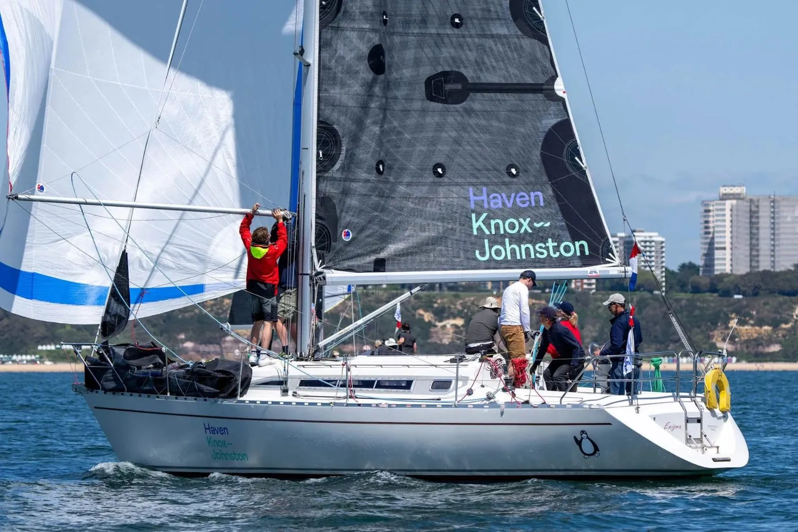 A group of sailors maneuvers a sailboat named 'Haven Knox Johnston' on a sunny day with buildings and a hillside in the background.