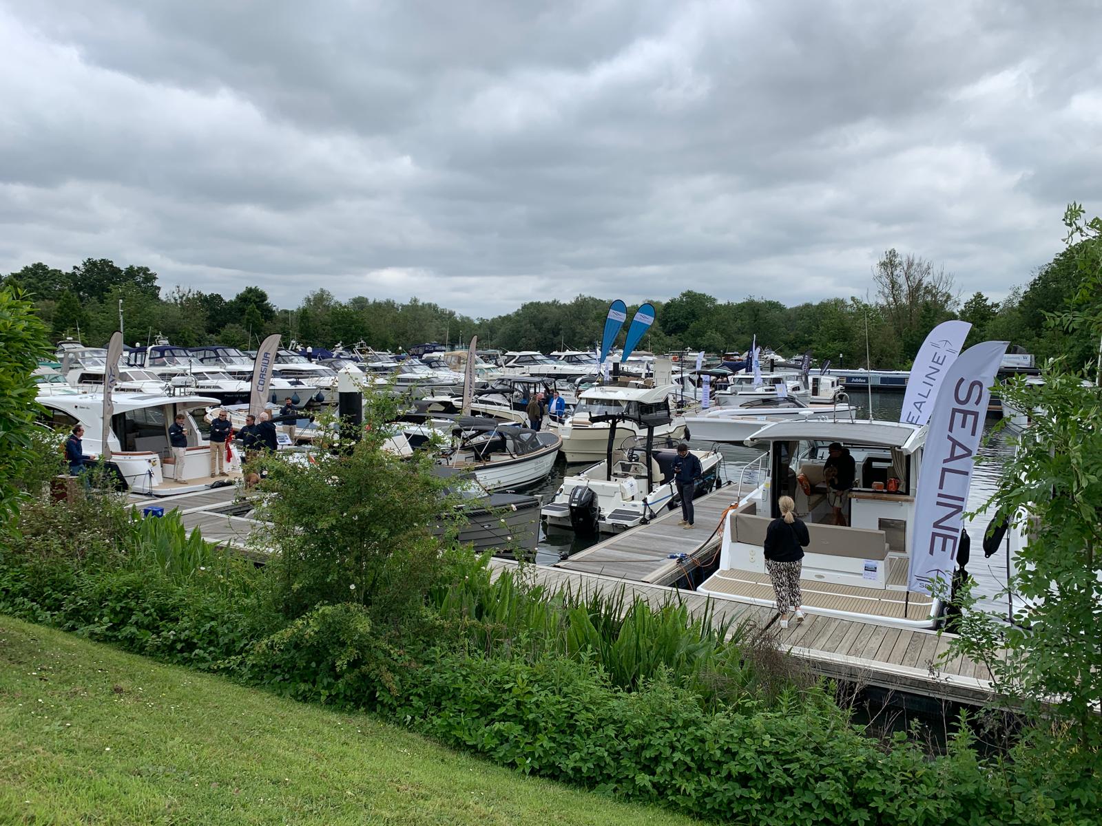 Numerous boats are docked at a marina with people walking along the boardwalks under a cloudy sky. Flags with branding are displayed.