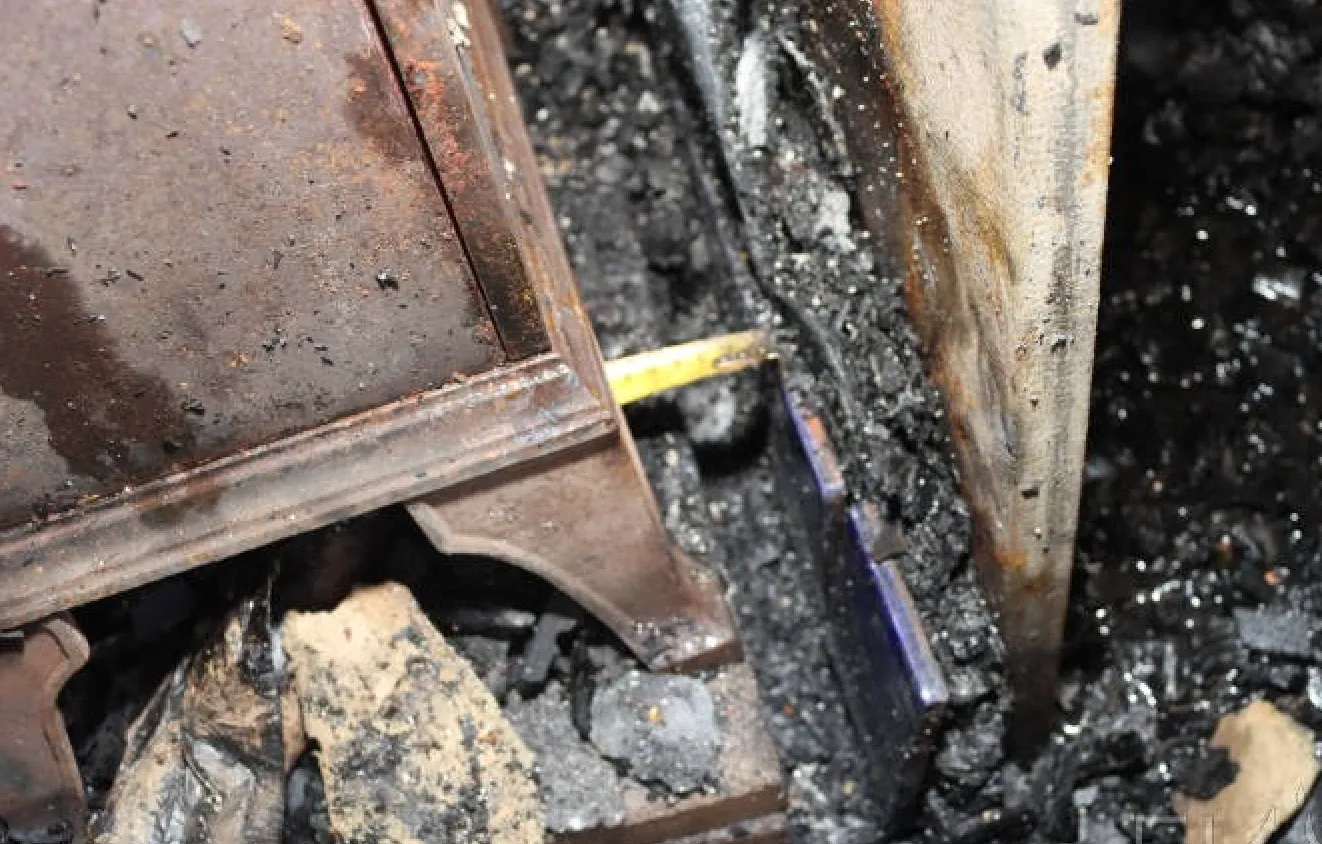 Close-up of a partially burned metal and wood structure with ashes and debris surrounding it.