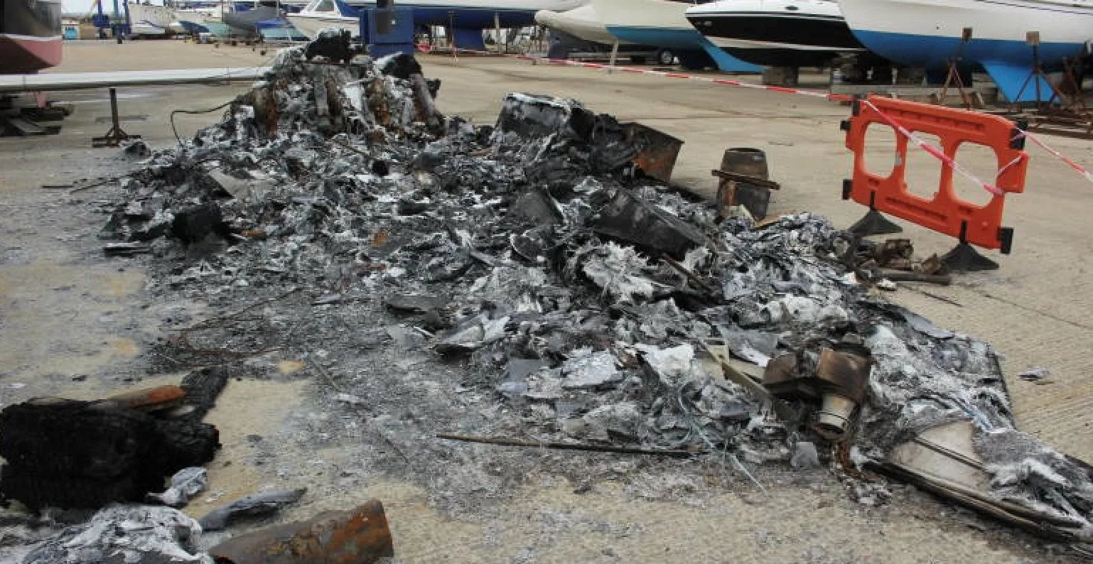 Burned debris of a boat remains on a concrete surface in a dockyard, with other boats in the background and barriers around the site.