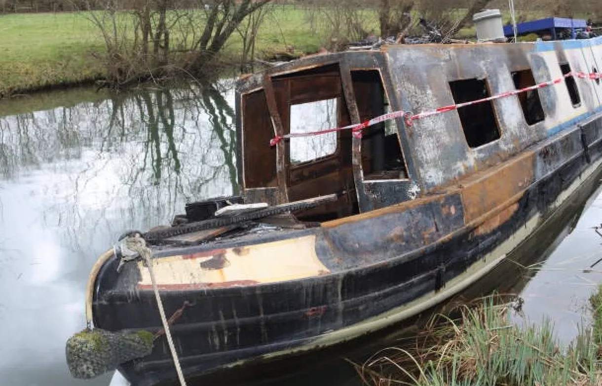 A partially burnt canal boat is taped off, lying in the water with visible fire damage, near a grassy and tree-lined bank.