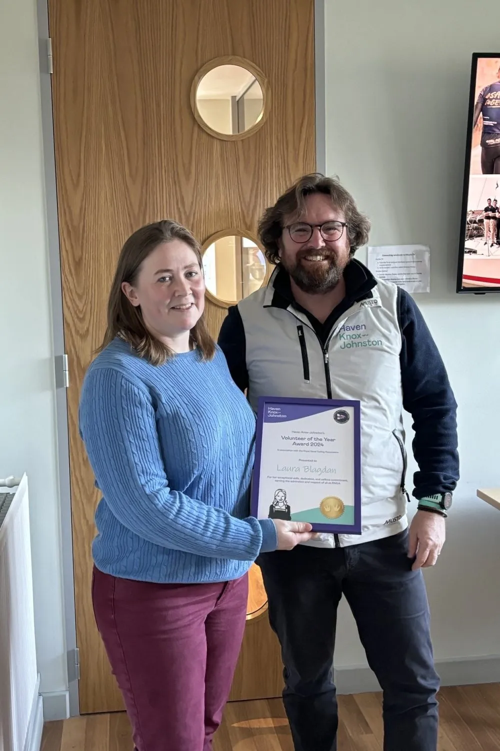 Two dedicated volunteers proudly hold an award certificate together, standing in a room with a wooden door as their backdrop.